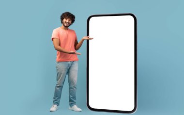 Şık, kıvırcık saçlı genç adam büyük cep telefonunun yanında duruyor. Mavi stüdyo arka planında beyaz ekran, boş ekran, model, kopyalama alanı, tam boy çekim alanı.