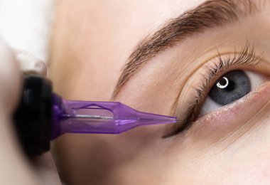 Mikro pigmentasyon kavramı. Güzellik uzmanı genç bir kadına kalıcı göz kalemi prosedürü uyguluyor. Çekici bir kadının güzellik stüdyosunda tedavi edilmesine yakın çekim yapıyor. Dövme makinesi kullanan bir sanatçı.