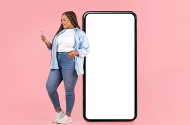 Pembe Stüdyo 'nun arka planında duran, boş ekran mesajlaşması ve akıllı telefon uygulaması ile büyük telefona yaslanan Afrikalı Amerikalı kadın. Mobil İletişim. Model