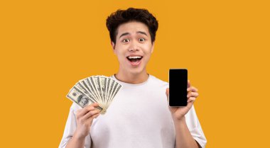 Elinde siyah boş ekranlı cep telefonu olan Asyalı bir adam elinde dolar banknotlarıyla nakit para hayranı turuncu arka planda yalnız poz veriyor. İnsanlar ve finans kavramı. Kopyalama alanı oluştur