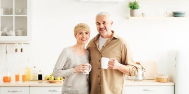 Romantik bir sabah, mutlu bir ilişki ve evde kahve içen yetişkin bir çift. Gülümseyen son sınıf öğrencisi ve sıcak çay bardaklarıyla minimalist beyaz mutfak iç mekanında kameraya bakan kadın ve erkek.