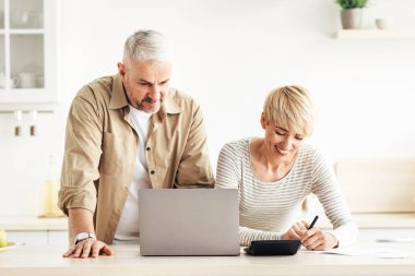 Ev muhasebesi, fatura ödeme ve planlama bütçesi, iyi haberler okumak, geri ödeme ya da ipotek onayı. Gülümseyen yetişkin kadın ve erkek dizüstü bilgisayara ve hesap makinesine bakar, mutfaktaki mali durumları kontrol eder.