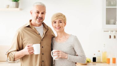 Birlikte mutlu sabahlar, kahve molası ve sabah içkisi. Covid sırasında evde güvende ol. Neşeli yetişkin erkek ve dişi ellerinde bir fincan sıcak çayla minimalist beyaz mutfak kameralarına bakıyorlar.