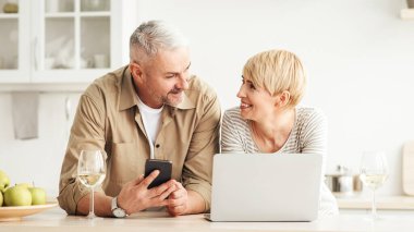 Teknoloji ve sosyal medyada boş zamanı olan orta yaşlı bir çift. Gülümseyen yetişkin erkek ve dişi birbirlerine modern mutfağın içinde bakıyorlar. Meyve ve ve kadehler dolusu şarap ve dizüstü bilgisayarla ve telefonla.