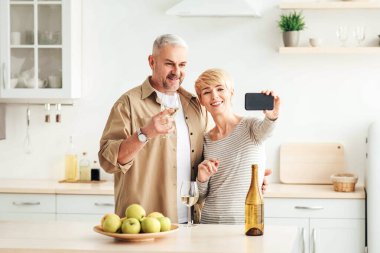 Hatırlanması gereken romantik anlar, kilitlenme sırasında evde buluşmalar, kendini tecrit etme, yeni normal şeyler. Neşeli, şarap kadehleri olan son sınıf ailesi minimalist mutfağın içinde meyvelerle akıllı telefondan fotoğraf çekiyor.