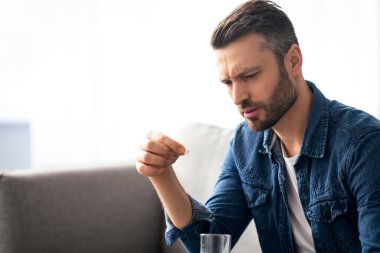 Tedavi, ilaç, ilaç, hap, vitamin, takviye konsepti. Bir bardak suyla ilaç alan üzgün sakallı bir adam. Orta yaşlı bir adamın tıpla yakın çekimi, baş ağrısı, kopyalama alanı