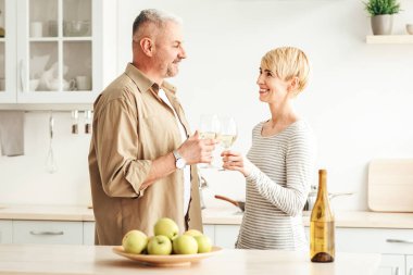 Yıl dönümünü, bayramı, doğum gününü ve sevgililer gününü evde, karantina sırasında kutlayın. Şaraplı mutlu orta yaşlı adam ve kadın, sıcak mutfak içlerine bardakları tokuşturur, boş alan