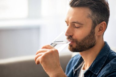 Sağlıklı yaşam tarzı, su içmesi, hidrasyon konsepti. Orta yaşlı, sakallı, bir bardak su içen, evinde taze su içen, fotokopi alanı olan, kenar manzaralı sağlıklı bir adam.
