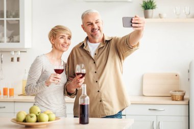 Evde bir randevu, kutlama, aile için selfie ve rahat bir daire. Mutlu yetişkin karı koca kırmızı şaraplı bardakları tutuyor ve modern mutfak iç mekanlarında sosyal ağlar için fotoğraf çekiyor.