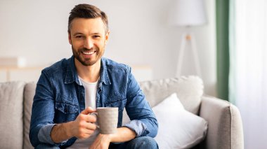 Bir fincan çayla kanepede oturan orta yaşlı, rahat oturma odasında kahve içen, kameraya gülümseyen, uzay fotokopisi çeken, beyaz sakallı bir adam.