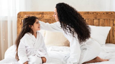 Küçük kızını seven genç bir anne evde birlikte otururken uzun saçlarını tarıyor. Güzel genç kadın ebeveyn çocuğuna bakıyor kıvırcık saçlı, ahşap fırçayla tarıyor.