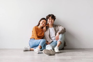 Yerde oturmuş beyaz stüdyo duvarının karşısında kameraya bakan sıradan kıyafetli neşeli, genç Asyalı bir çiftin tam boy portresi. Milenyum eşleri birlikte poz veriyor ve gülümsüyor.