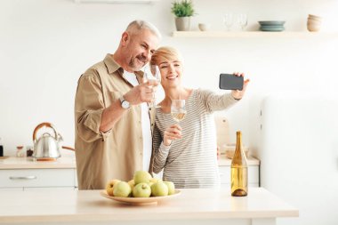 İnternette, video görüşmesinde ve covid-19 sırasında tatilde tebrik ederim. Hafif şaraplı mutlu Avrupalı çift minimalist mutfağın içinde, boş bir alanda akıllı telefondan fotoğraf çekiyor.