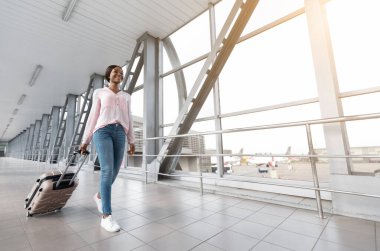 Havaalanı terminalinde bavuluyla yürüyen mutlu Afrikalı Amerikalı kadın portresi, neşeli siyah kadın iş gezisi veya yaz tatiline hazır, kopyalama alanı.
