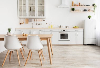 İskandinav tarzı hafif mutfak tasarımı. Ahşap yemek masası ve raflarda farklı mutfak aletleri ve aksesuarları olan beyaz mobilyalar, pencerenin yanındaki küçük beyaz buzdolabı, boş alan.