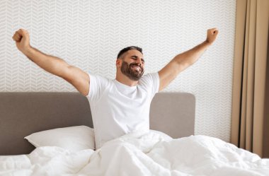 Avrupalı sakallı bir adam modern yatak odasında uyanıyor ve kollarını mutlu bir gülümsemeyle açıyor, beyaz çarşaflarla yatakta oturuyor. Ev konseptinde rahat bir sabah rutini.