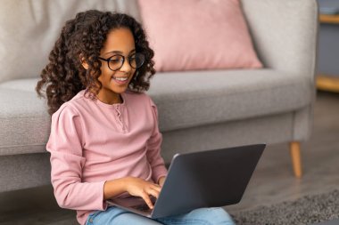 E-öğrenme konsepti. Afro-Amerikalı kız dizüstü bilgisayarını kullanıyor, halının üzerinde oturuyor, kanepeye yaslanıyor, klavyeye yazı yazıyor. Öğrenci kız ev ödevi yapıyor, evde online kurs izliyor.