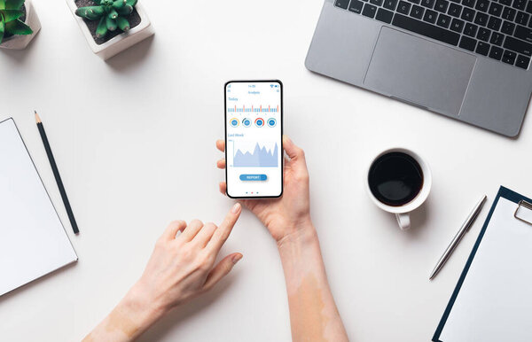 Female hands holding smartphone with analytics app, white workdesk with laptop, cup of coffee and plant background, top view. Cropped of business analyst using mobile phone, creative image