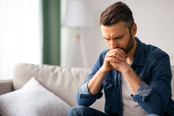 Расстроенный бородатый мужчина среднего возраста в повседневной одежде сидит на диване в гостиной, думая о чем-то, глядя вниз, разочарованный человек, имеющий финансовые проблемы во время пандемии, копировать пространство