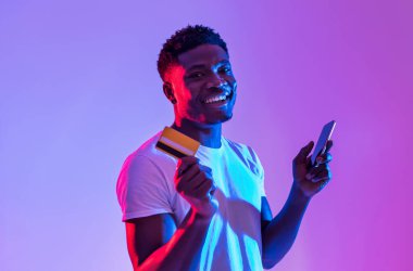 Portrait of cheerful young black man holding cellphone and credit card, using banking or shopping mobile app, receiving cashback, trading or gambling online in neon light