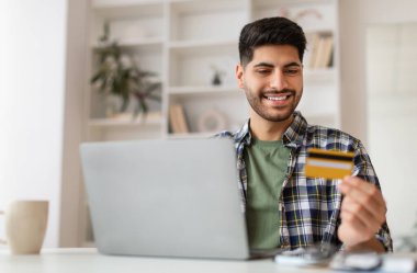 Kolay Ödeme ve Nakit Konsepti. Elinde kredi kartı tutan heyecanlı milenyum adamının portresi ve evde oturup web mağazasından alım-satım siparişi veren finansal işlemler yapan bilgisayarını kullanması.