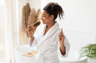 Mutlu siyah kadın saç fırçasıyla şarkı söylüyor ve evde modern banyoda beyaz bornoz giyerek saç bakımı rutininin tadını çıkarıyor..