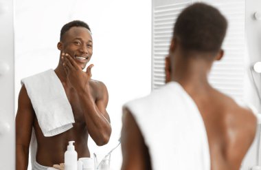 Tıraşlı gömleksiz Afrikalı Adam, sabah tıraş olduktan sonra pürüzsüz yüzüne dokunuyor, aynaya bakıyor, modern banyoda evde duruyor. Erkek Yüz Teni Rutin Konsepti
