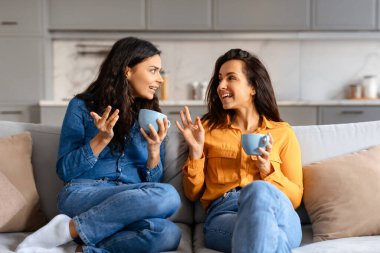 Sıcak bir sohbete tutulan iki genç kadın, evdeki bej bir kanepede kahve içerken hikayelerini paylaşıyorlar.