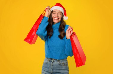 Mavi kazaklı ve Noel Baba şapkalı mutlu bir kadın elinde kırmızı alışveriş çantaları, sarı arka planda duruyor, bayramlara hazırlanıyor ve hediyeler alıyor. Noel alışverişi konsepti