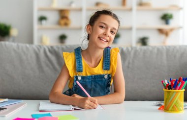 Modern Eğitim Konsepti. Oturma odasında oturan gülümseyen küçük kız öğrencinin portresi, defterine veya günlüğüne notlar yazması, kameraya poz vermesi, seçici odaklanma.