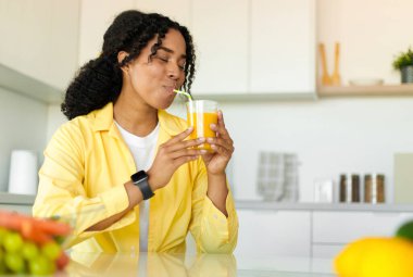 Young african american woman drinking fresh orange or mango juice from glass, sitting in kitchen, copy space. Happy lady enjoying delicious fruit drink. Detox and healthy nutrition concept