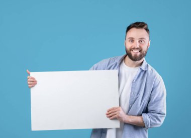 Neşeli beyaz adam mavi stüdyo arka planında beyaz boş plaket sergiliyor, tasarım için maket yapıyor. Boş reklam afişi olan mutlu hippi adam, tanıtımın için yer aç.