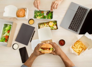 Break at work, lunch and healthy food delivery. Young woman eating salad with vegetables, man holding burger with meat substitute, at table with boxes of food, notepad and laptop, top view, copy space