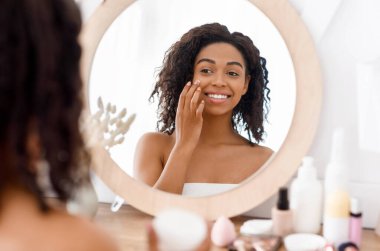 Güzel siyah kadın aynaya bakıyor ve yüz nemlendirici kremi sürüyor, havluya sarılıp banyoya sarılıyor, omuz hizasına seçici bir şekilde bakıyor.