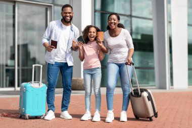 Sonunda tatil zamanı. Biletleri olan ve yumruklarını sıkmış üç kişilik neşeli, pozitif siyah aile, kazanan jest yapıyor, havaalanı terminalinin yanında birlikte duruyorlar.