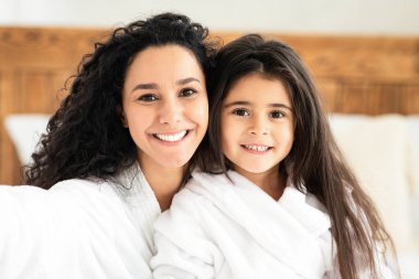 Beyaz bornozlu güzel bir anne ve kız çocuğu gülümsüyor ve yatak odasının içinde selfie çekiyor. Sevgi dolu ve neşeli esmer kadın neşeli küçük kızına sarılıyor, boşluğu kopyalıyor.