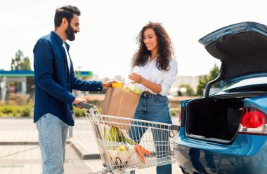 Evli orta doğulu çift arabaya sağlıklı yiyecek dolu kağıt torba koyuyor, bir adam karısına yardım ediyor, alışveriş merkezinin yakınındaki park yerinde dikiliyor.