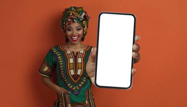 Mutlu, neşeli, oldukça geleneksel Afrikalı bir kadın elinde kocaman, beyaz ekranlı akıllı bir telefon tutuyor, siyah kadın ulusal kostüm giyiyor, turuncu stüdyo geçmişi var, model, afiş.