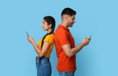 Sırt sırta verip akıllı telefonlar kullanan neşeli orta doğulu çift, mobil oyunlar oynayan mutlu Arap erkek ve kadın ya da mavi arka planda poz verirken internetten mesajlaşıyorlar.