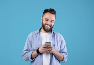 Mavi stüdyo arka planında akıllı telefon kullanan beyaz bir gencin portresi. Modern aletlerle gülümseyen milenyum adamı internette sohbet ediyor, internette geziniyor, sosyal medyada paylaşıyor.