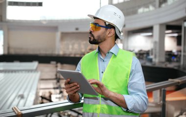 Bin yıllık ciddi bir İslami mühendis koruyucu üniformalı, gözlüklü ve tablet kontrollü sakallı sert şapkalı fabrika işçisi. İş ve endüstri covid-19 salgını nedeniyle gelişiyor