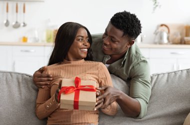 Afro-Amerikan erkeğinin karısına sarılması, hediye kutusu vermesi, Sevgililer Günü için büyük bir sürpriz hazırlaması, evin içi, boş yer. Mutlu siyah adam kız arkadaşını kucaklıyor, hediyesini tutuyor.