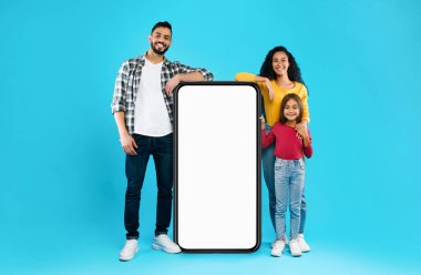 Mobil Uygulama Reklamları İçin Boş Ekran Arkasında Mavi Stüdyo Poz Veren Büyük Telefon Yanındaki Arap Aile. Arap Ebeveynler ve Kız Reklam Uygulaması. Model