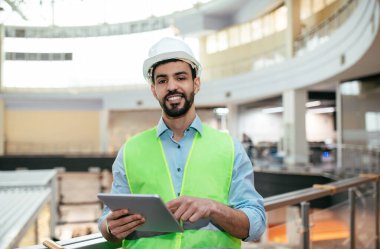 Sakallı, yakışıklı, genç, Arap bir mühendis. Fabrikanın iç kısımlarında tablet işi olan koruyucu üniformalı. Modern cihaz, fabrikada profesyonel, genel mimar kontrol endüstrisi