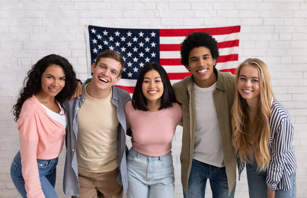 Fun education, language courses, learn english and exchange students. Young smiling people of different nationalities hug in college with large USA flag on white brick wall, free space, studio shot