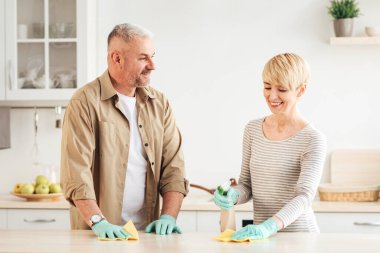 Ev işleri, ev işleri ve evdeki izolasyon sırasında temizlik. Lastik eldivenli mutlu erkek ve kadın minimalist mutfak iç dekorasyonunda sprey ve süngerle masayı siliyor.