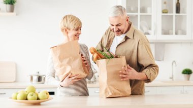 Market alışverişi, birlikte ev işleri, Cvid-19 tecrit sırasında online yemek teslimatı. Gülümseyen orta yaşlı kadın ve erkek mutfak içi sebzeli eko kağıt torbalarla dolu boş bir yer.
