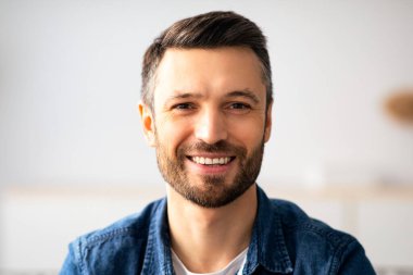 Mutlu, sakallı, sıradan kıyafetli, ev içinde çekilmiş yakın plan bir portre. Keyifli orta yaşlı bir adam rahat bir oturma odasında dinleniyor, kameraya gülümsüyor, mekanı kopyalıyor.