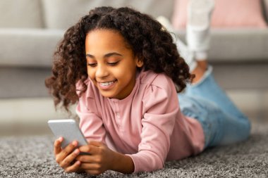 Dijital yerliler. Afro-Afrikalı Amerikalı genç kız akıllı telefonunu kullanıyor, sosyal medyada sohbet ediyor ve mesajlaşıyor, dinleniyor ve oturma odasındaki halıya uzanıyor.