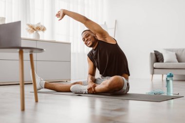 Çevrimiçi Eğitim. Cheerful Black Man, evde jimnastik yaparken dizüstü bilgisayarda esneme egzersizi yapıyor. Atletik Afrikalı Amerikalı Adam Oturma odasında Yerde Egzersiz Yapıyor
